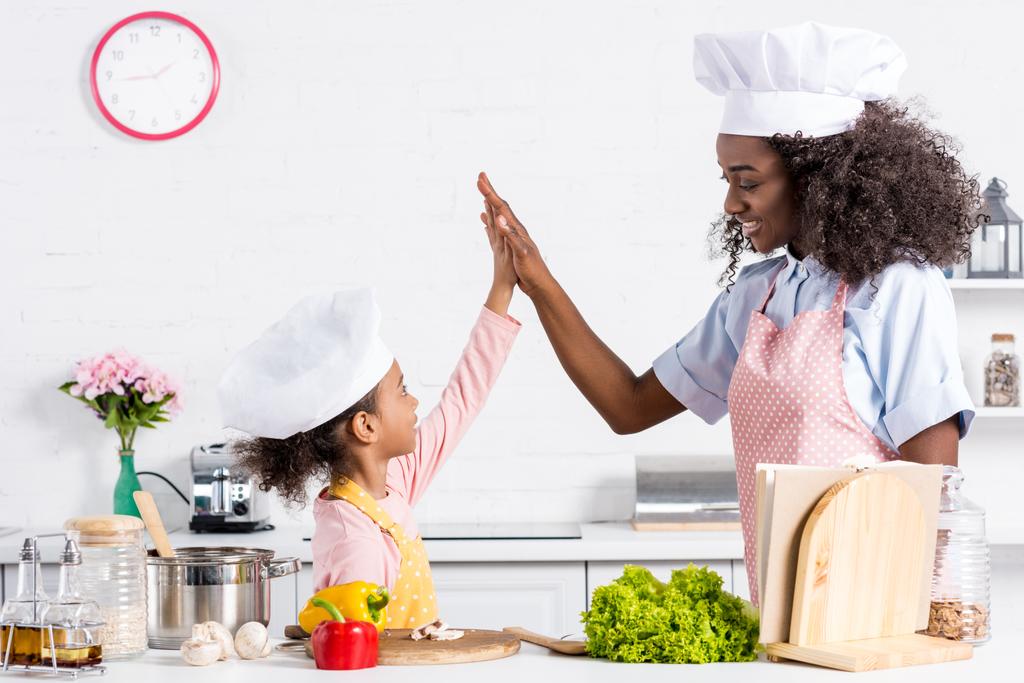 african american mother and daughter in chef hats cooking and giving high five on kitchen - Photo, Image