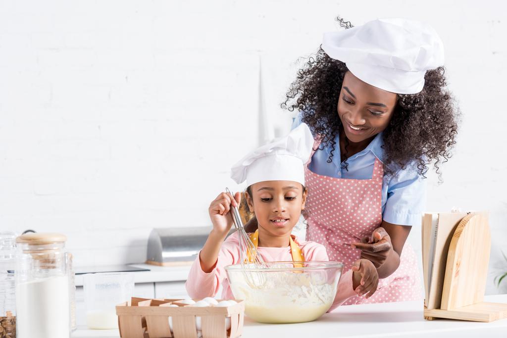 Αφρικανική αμερικανική μητέρα και κόρη στην καπέλα σεφ ανάμειξη ζύμη στην κουζίνα με βιβλίο μαγειρικής - Φωτογραφία, εικόνα