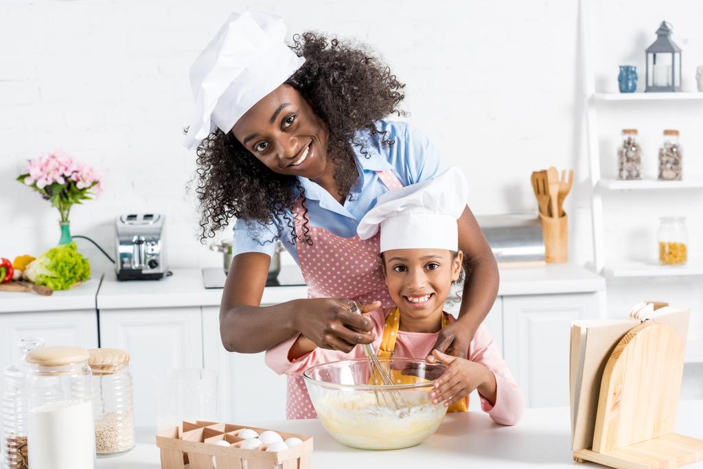 キッチンに泡立て器で生地を混合シェフ帽子でアフリカ系アメリカ人の母と娘 - 写真・画像