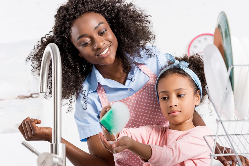 heureux afro-américain mère et fille laver la vaisselle avec éponge et mousse sur la cuisine
 - Photo, image