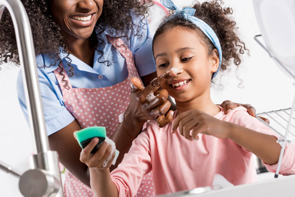 Afrikanisch-amerikanische Mutter und Tochter haben Spaß beim Geschirrspülen mit Schwamm in der Küche - Foto, Bild
