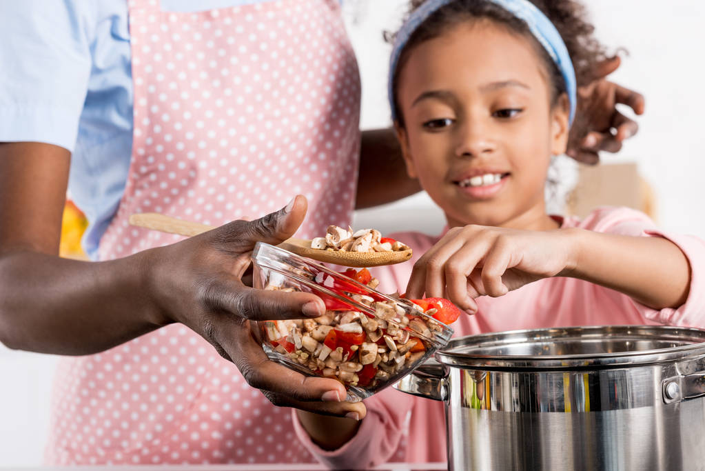 afro-americana madre e figlia mettendo ingredienti in casseruola in cucina
  - Foto, immagini