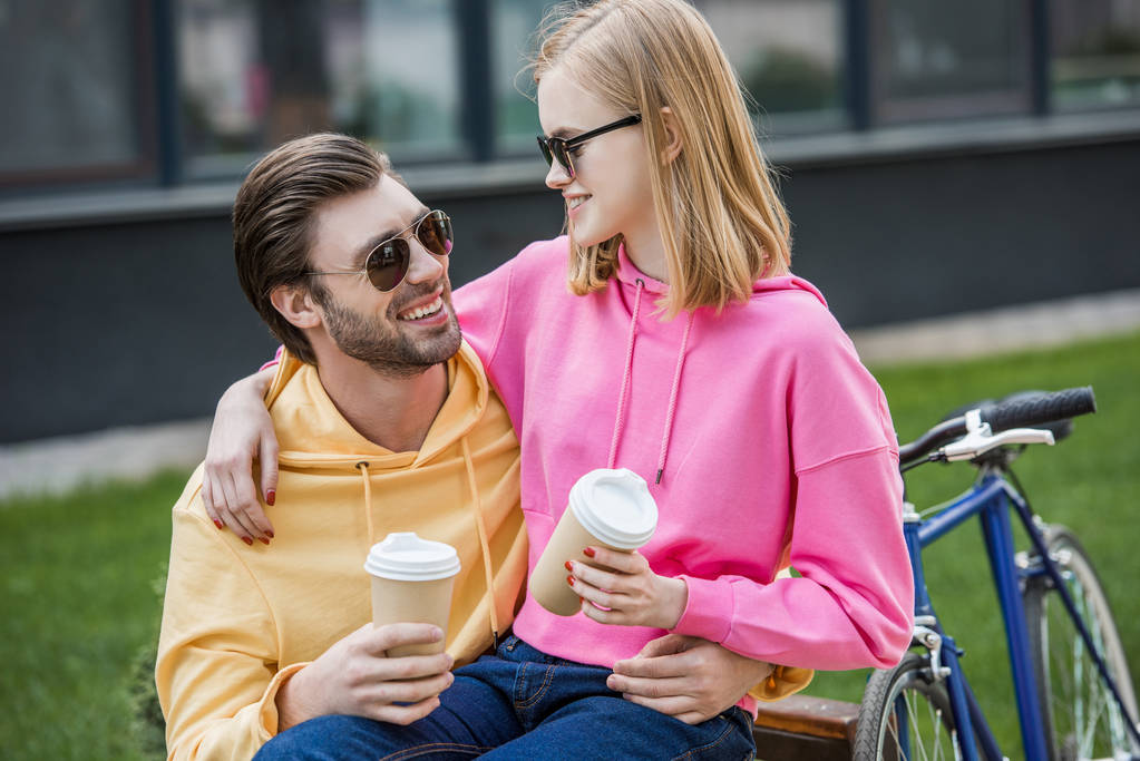 smiling stylish couple in sunglasses sitting on bench with papers cups of coffee  - Photo, Image
