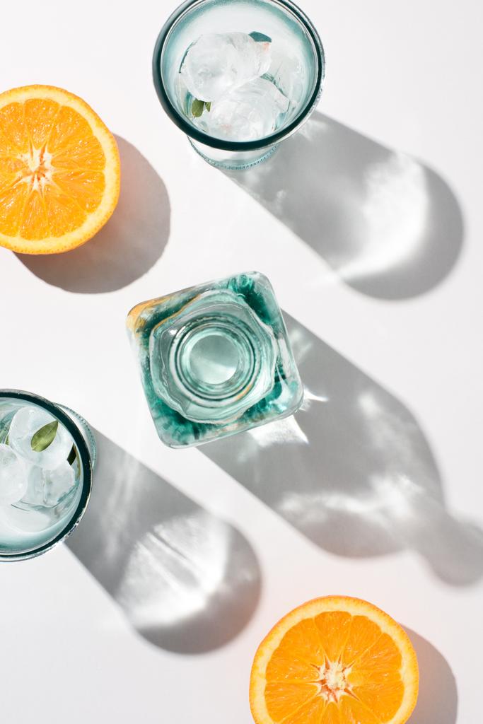 Ansicht von Orangenstücken, Flaschen und Gläsern mit Eis auf weißer Oberfläche - Foto, Bild