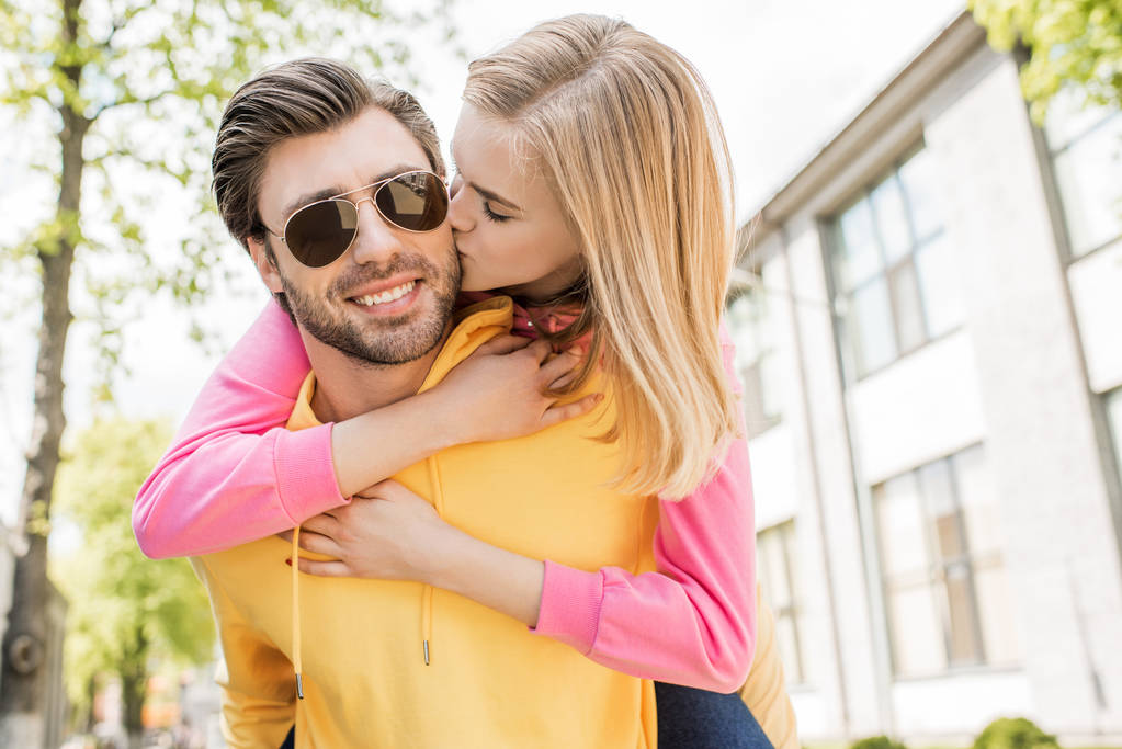 hombre con estilo en gafas de sol haciendo paseo a cuestas a su novia mientras ella lo besa en la mejilla
 - Foto, imagen