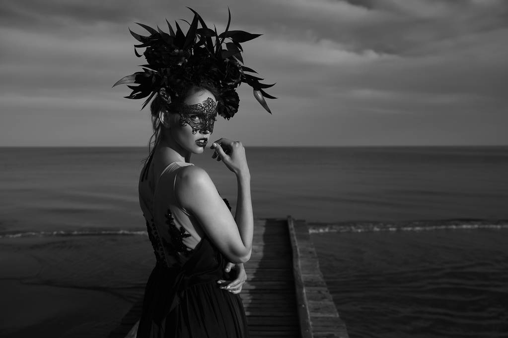 Black and white dramatic portrait of beautiful emotional woman with black lace mask on face and big floral crown on head stands on wooden pier. Dark clouds and sea on background - Photo, Image
