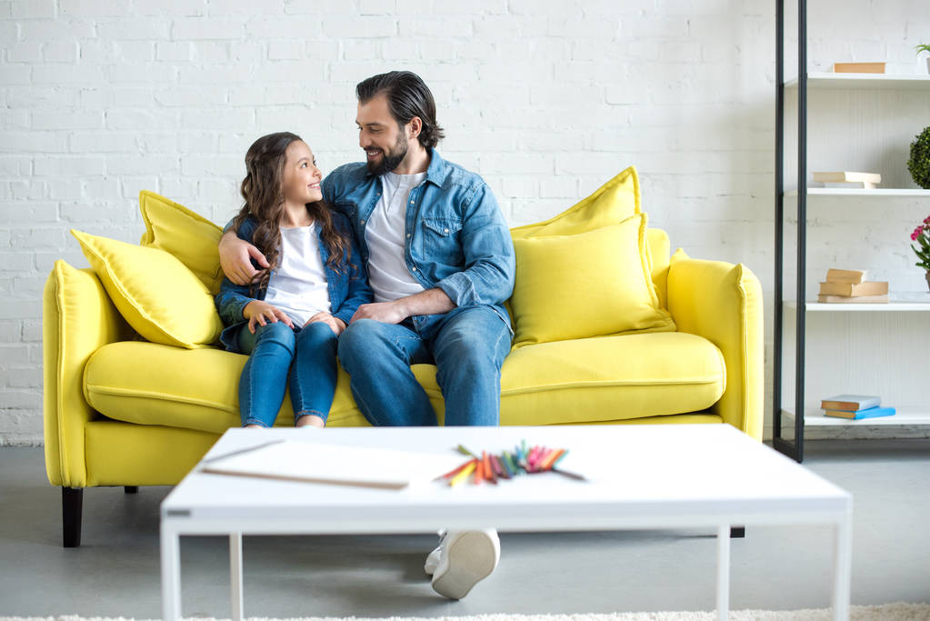счастливые отец и дочь сидят вместе на желтом диване и улыбаются друг другу дома
 - Фото, изображение