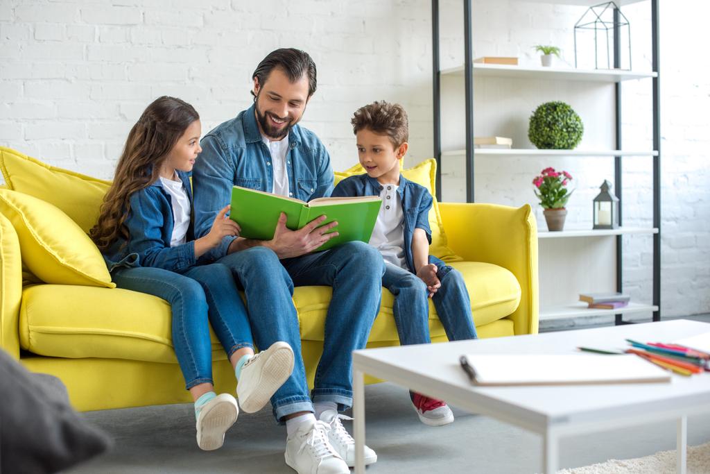 Lächelnder Vater mit Kindern beim gemeinsamen Lesen auf dem heimischen Sofa  - Foto, Bild