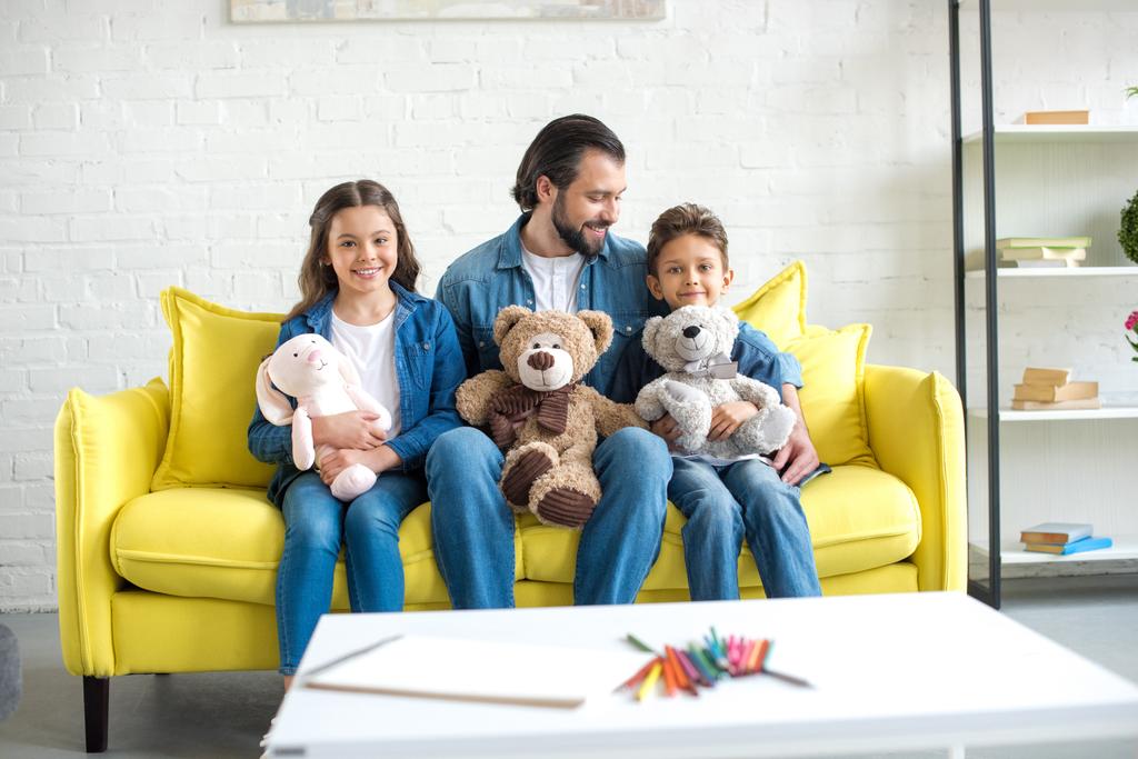 счастливый отец с очаровательными маленькими детьми, держащими игрушки и сидящими дома на диване
 - Фото, изображение