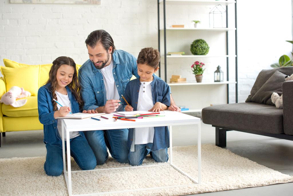glücklicher Vater mit zwei Kindern, die auf Teppich knien und zu Hause mit Buntstiften zeichnen - Foto, Bild