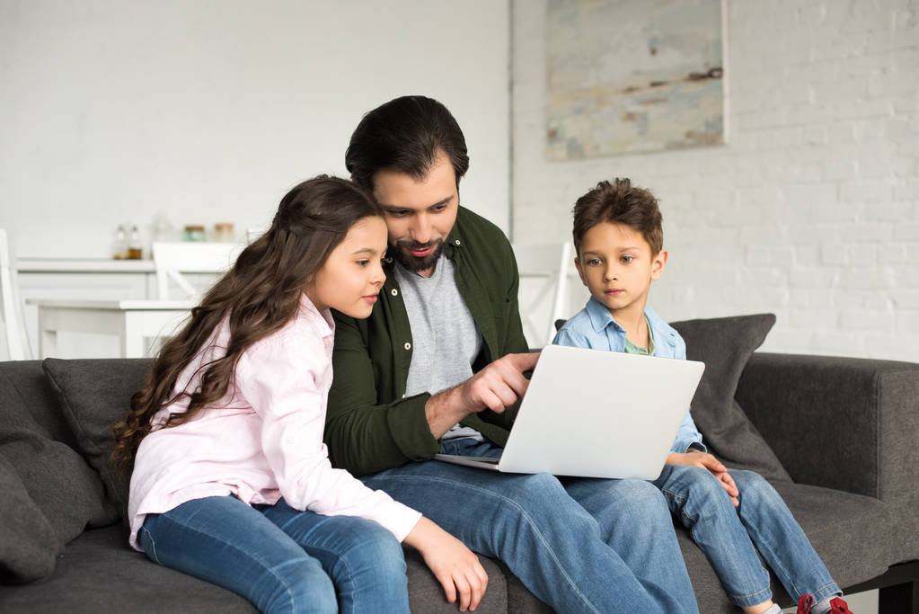 apa és két aranyos gyerek ül a kanapén, és laptop együttes használata  - Fotó, kép