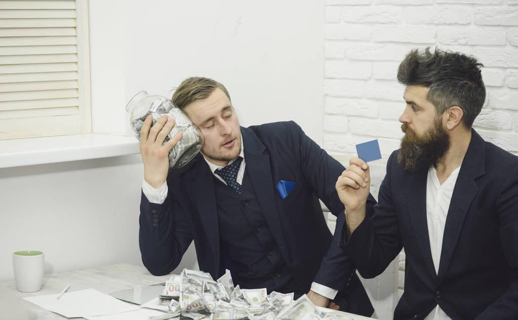 Tired man holds jar full of cash, colleague holds credit card. Business partners, businessmen at meeting in office. Business profit, keeping money in jar or bank account. Cash and banking concept. - Photo, Image