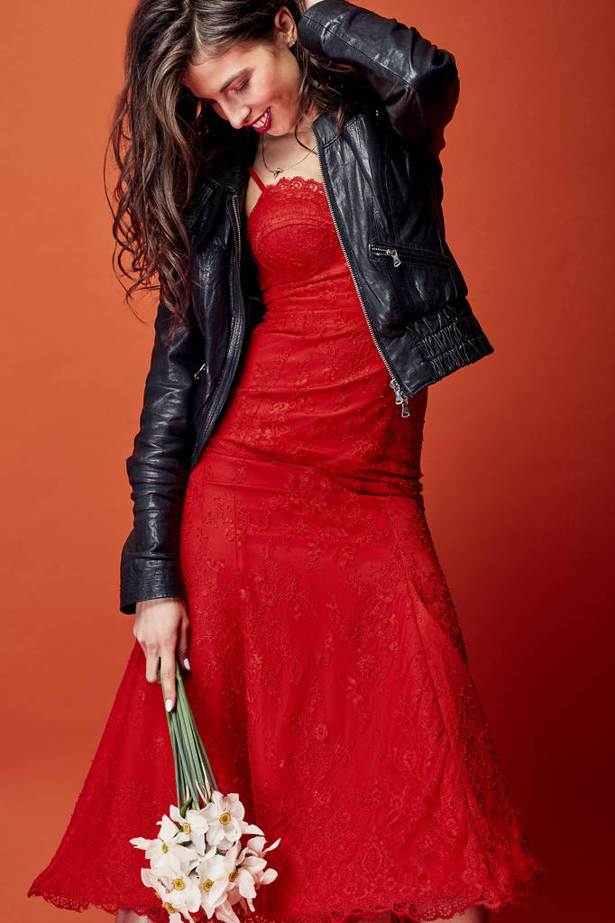 ウェーブのかかった髪と美しい笑顔ブルネットの女性服を着た赤レース ドレス、黒革のジャケットがオレンジ色の背景とスタジオで水仙の束でポーズ  - 写真・画像