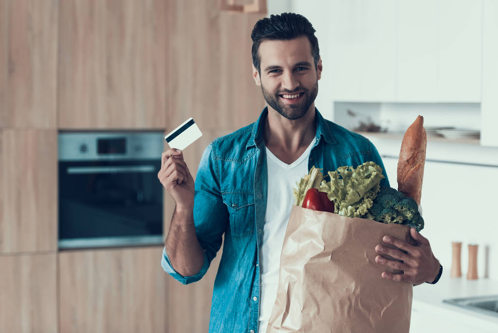 Handsome man with credit card and package of products is standing in kitchen. - Photo, Image