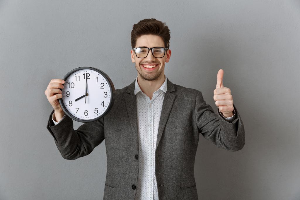 muotokuva hymyilevä liikemies kello kädessä osoittaa peukalo ylös harmaalla seinällä tausta
 - Valokuva, kuva