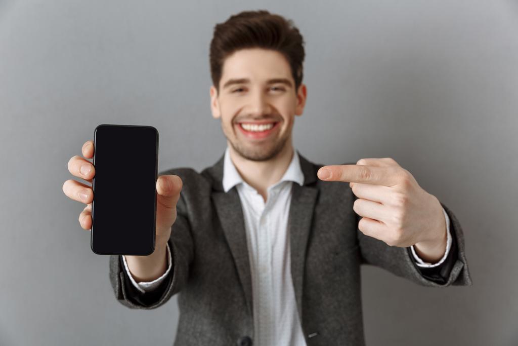Selektywny fokus uśmiechający się biznesmen, wskazując na smartphone rezygnować pusty ekran w ręku na tle szarej ścianie - Zdjęcie, obraz