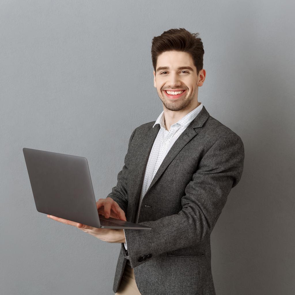 χαμογελώντας επιχειρηματία στο κοστούμι με φορητό υπολογιστή φόντο γκρίζο τοίχο - Φωτογραφία, εικόνα