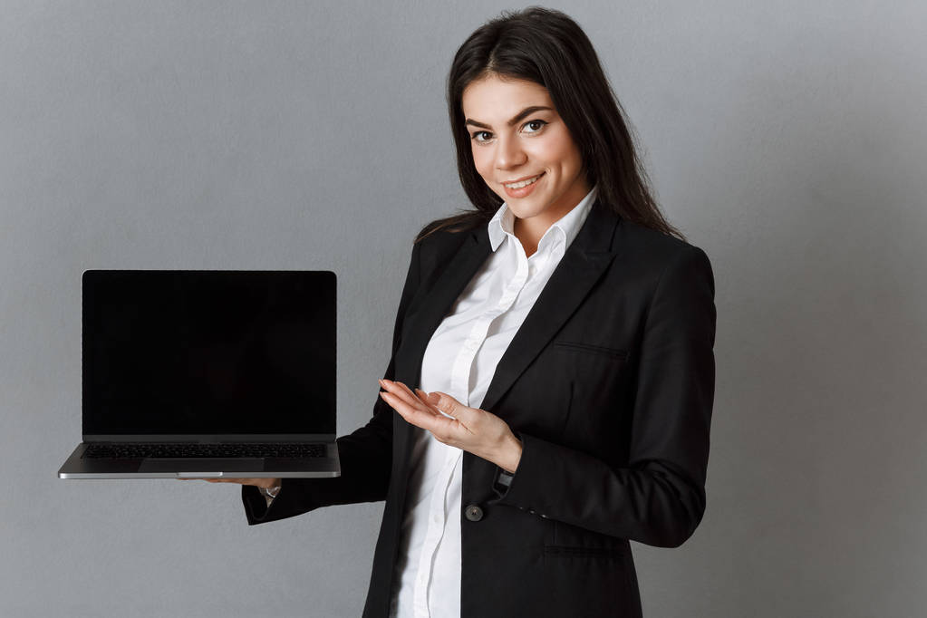 улыбающаяся деловая женщина, указывающая на ноутбук с чистым экраном на фоне серой стены
 - Фото, изображение