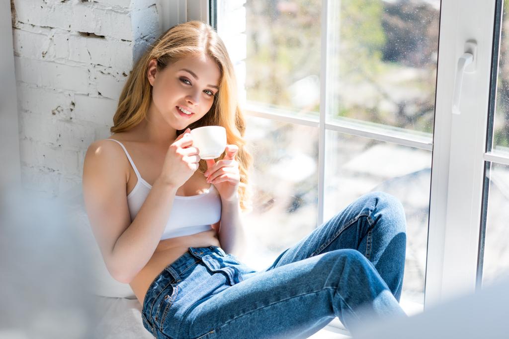 ευτυχής όμορφη κορίτσι που κάθεται στο περβάζι με φλιτζάνι καφέ - Φωτογραφία, εικόνα