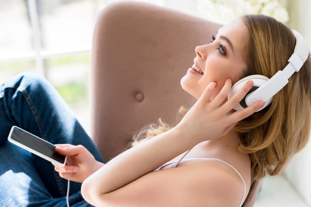 улыбающаяся молодая женщина слушает музыку с наушниками и смартфоном на кровати
 - Фото, изображение