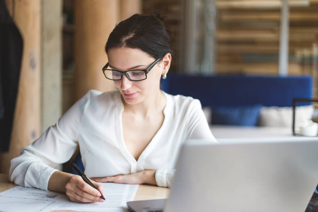 Brunette Femme d'affaires avec des lunettes travaillant derrière un ordinateur portable et avec des papiers près de la fenêtre dans une atmosphère de café confortable
 - Photo, image