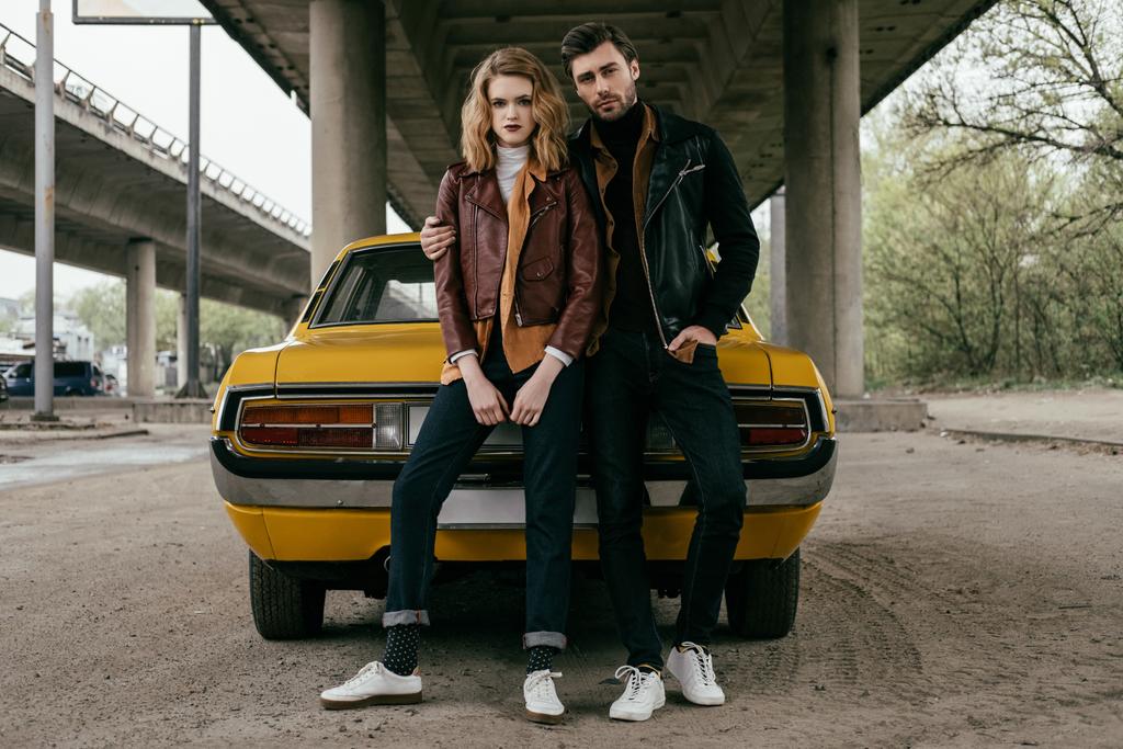 full length view of young couple in leather jackets sitting on yellow old-fashioned car and looking at camera - Photo, Image