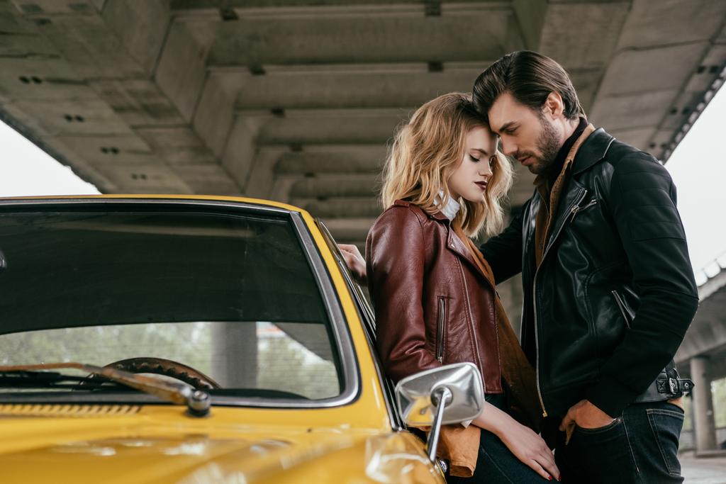 вид сбоку стильной молодой пары, обнимающейся возле желтого ретро-автомобиля под мостом
  - Фото, изображение