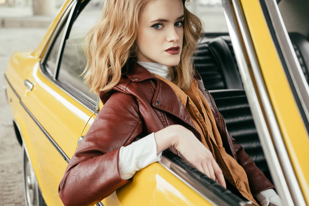 όμορφο κομψό κορίτσι με δερμάτινο μπουφάν συνεδρίαση στις παλιομοδίτικο αυτοκινήτου και βλέπουν τα φωτογραφικών μηχανών - Φωτογραφία, εικόνα