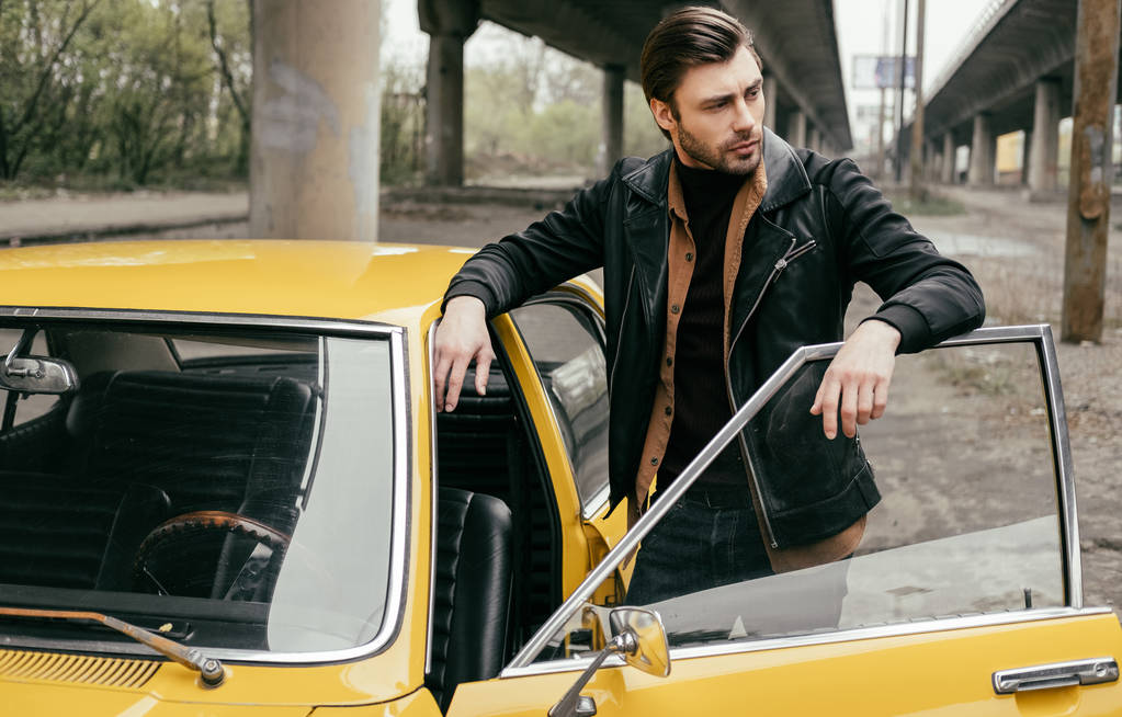 όμορφος μοντέρνος νεαρός άνδρας στο δερμάτινο μπουφάν στέκεται κοντά αυτοκίνητο ρετρό και αναζητούν μακριά - Φωτογραφία, εικόνα