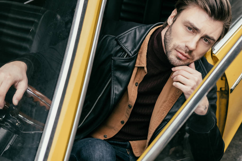 pensive handsome young man sitting in classic car and looking at camera - Photo, Image