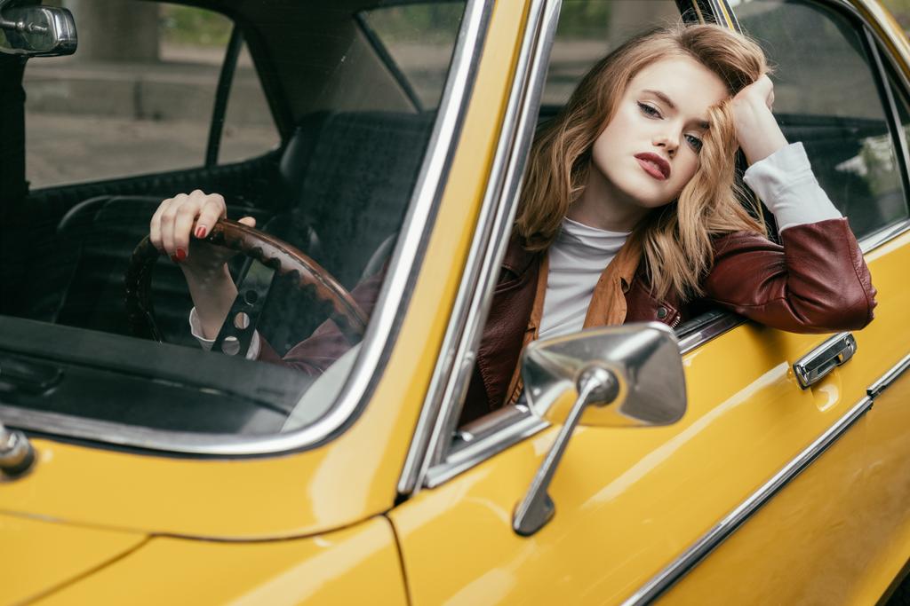 魅力的な若い女性はカメラ目線とステアリング ホイールを押しながらレトロな車に座っています。  - 写真・画像
