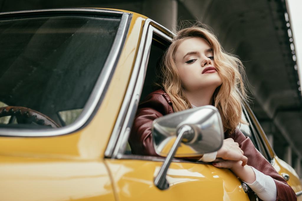 csábító lány látszó-on fényképezőgép-sárga retro autó ülve bőrkabát - Fotó, kép