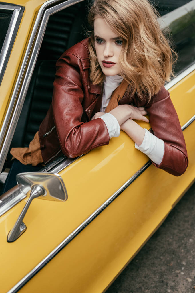 attractive girl in leather jacket looking through window of yellow vintage car - Photo, Image
