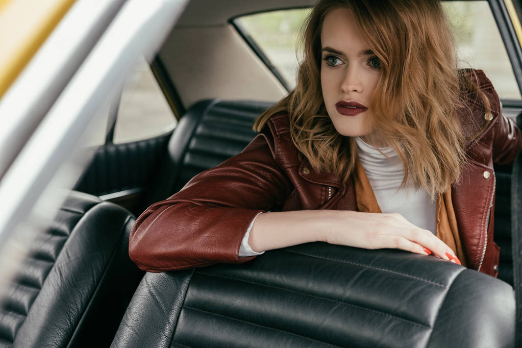 beautiful young woman in leather jacket sitting in classic car and looking away - Photo, Image