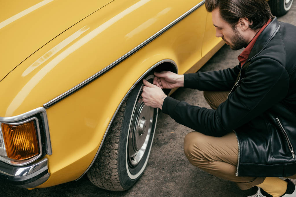 обрезанный снимок красивого мужчины в кожаной куртке, закрепляющий колесо жёлтого ретро-автомобиля
  - Фото, изображение
