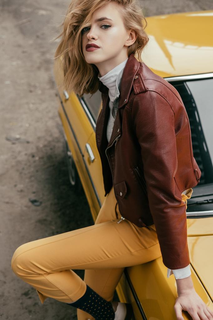 mujer joven con estilo en chaqueta de cuero mirando a la cámara mientras está sentado en el coche vintage amarillo
 - Foto, imagen