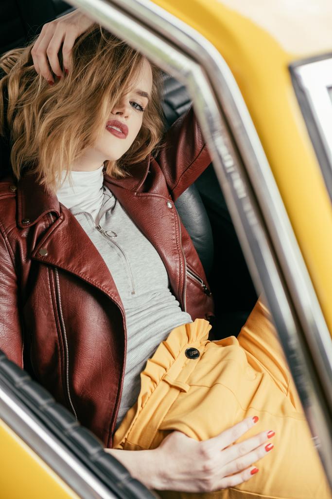 wysoki kąt widzenia stylowa dziewczyna w skórzanej kurtce siedzi w samochodzie rocznika i patrząc na kamery    - Zdjęcie, obraz