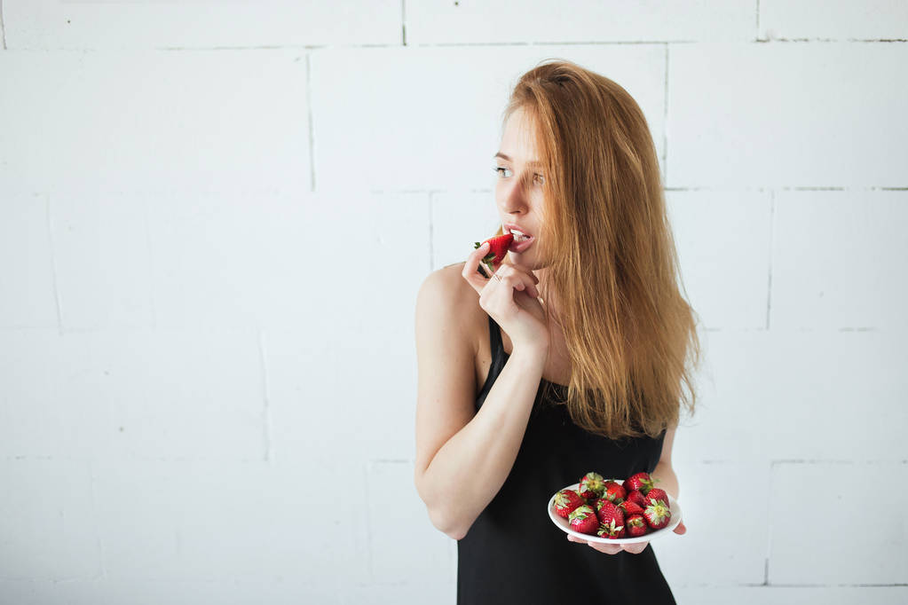 Mulher magra sexy nova comendo morango suculento doce perto da parede branca. Luz natural, espaço de cópia
 - Foto, Imagem