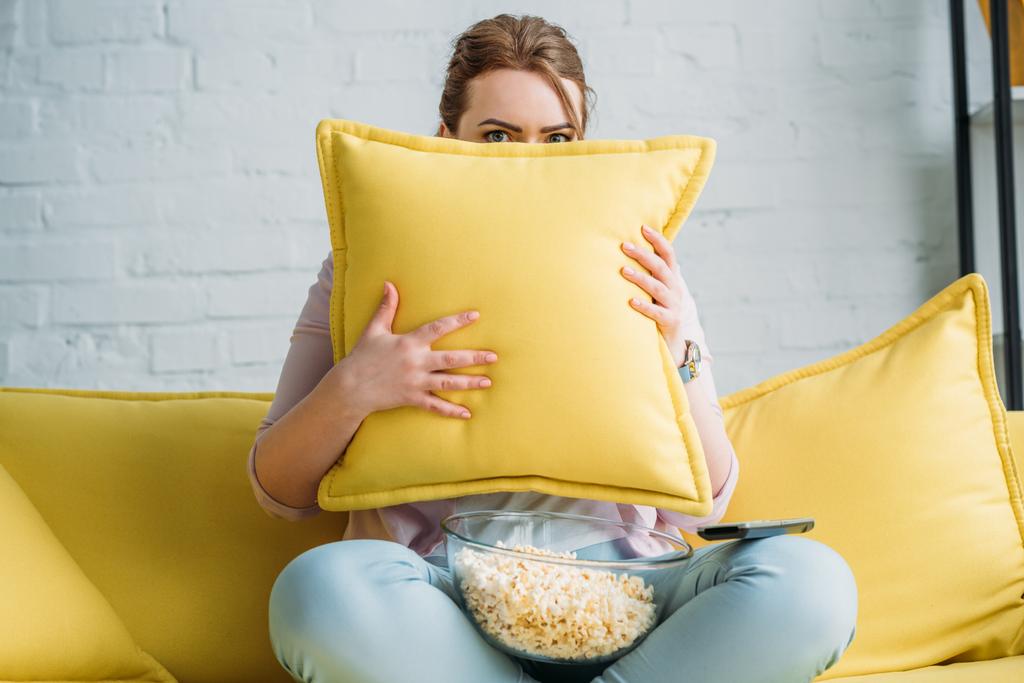 woman looking out from cushion while watching horror movie with popcorn at home - Photo, Image