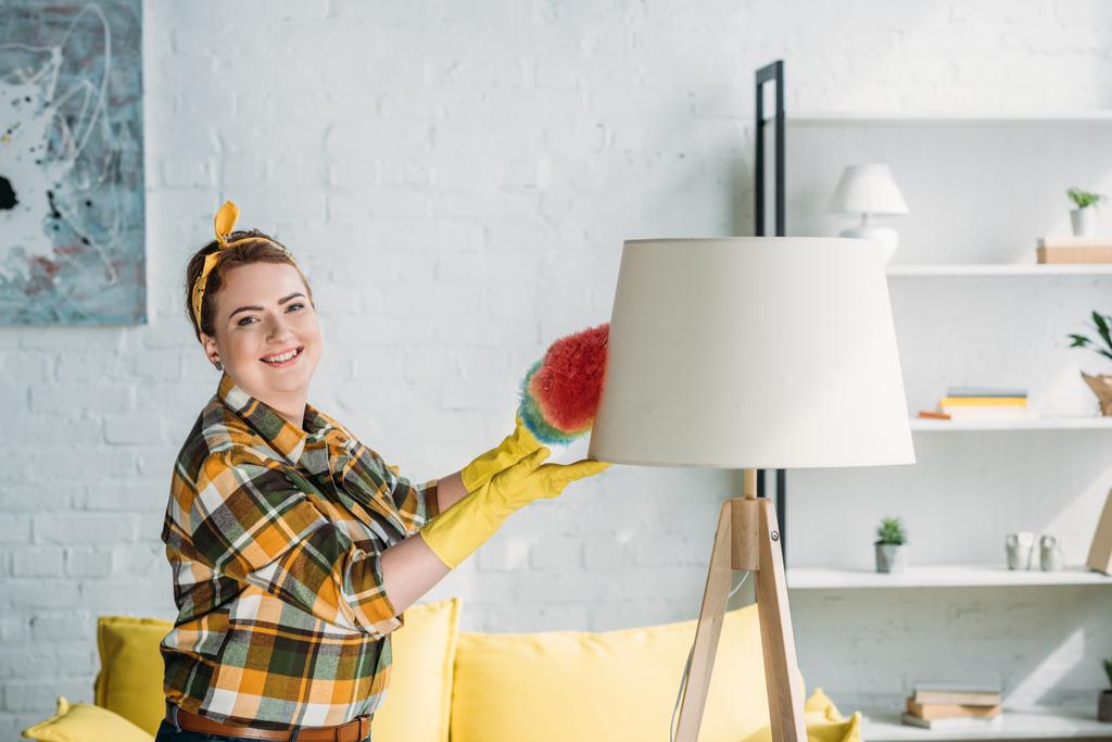 mooie vrouw afstoffen lamp met stof borstel thuis - Foto, afbeelding