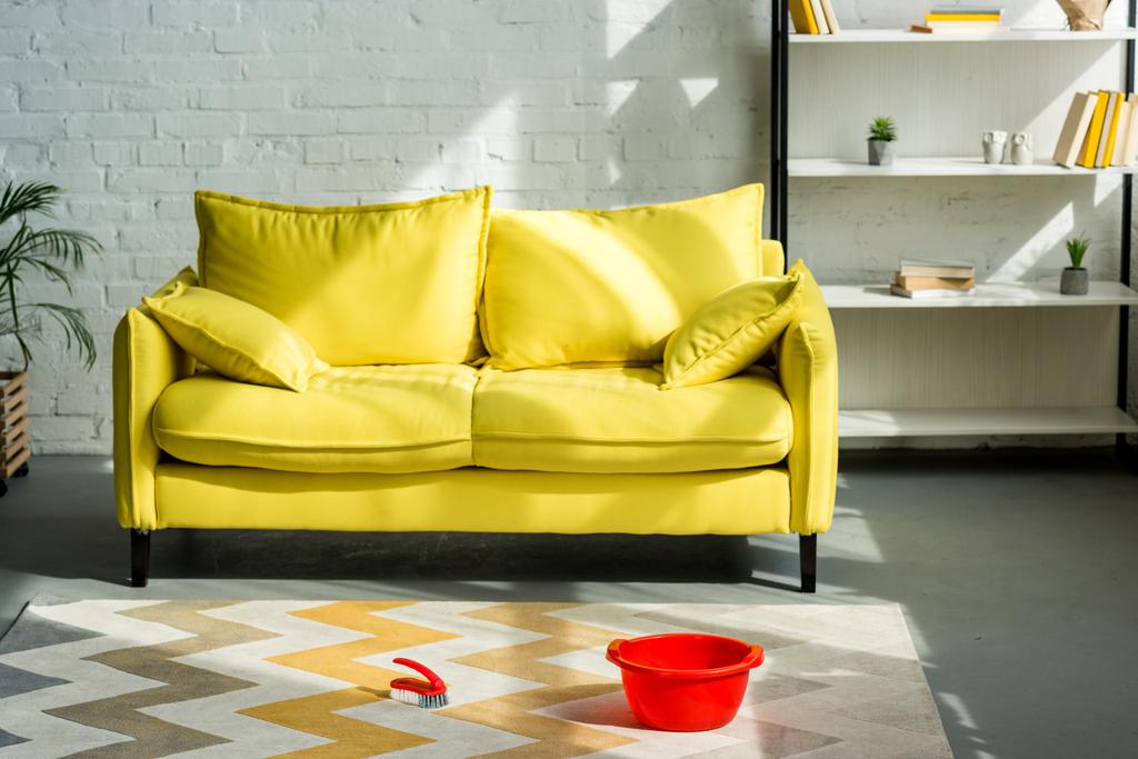 κόκκινο κουβά και βούρτσα καθαρισμού σε χαλί στο πάτωμα, κίτρινος καναπές στο σαλόνι - Φωτογραφία, εικόνα