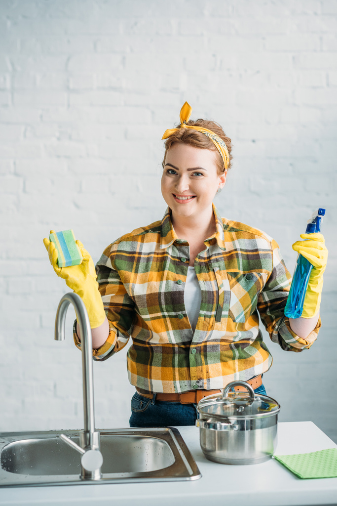 χαμογελαστό γυναίκα κρατώντας πλύσιμο σφουγγαριών και σπρέι μπουκάλι κατά τον καθαρισμό στην κουζίνα - Φωτογραφία, εικόνα