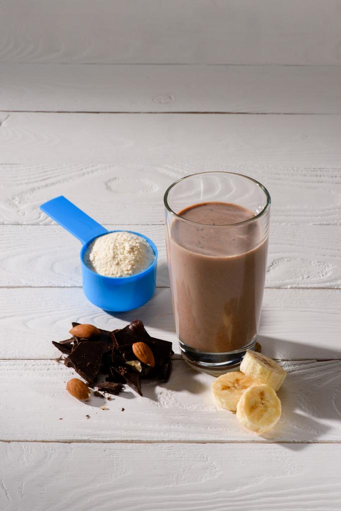 Бокал протеинового коктейля с миндалем, бананом и шоколадом на белом деревянном столе
 - Фото, изображение