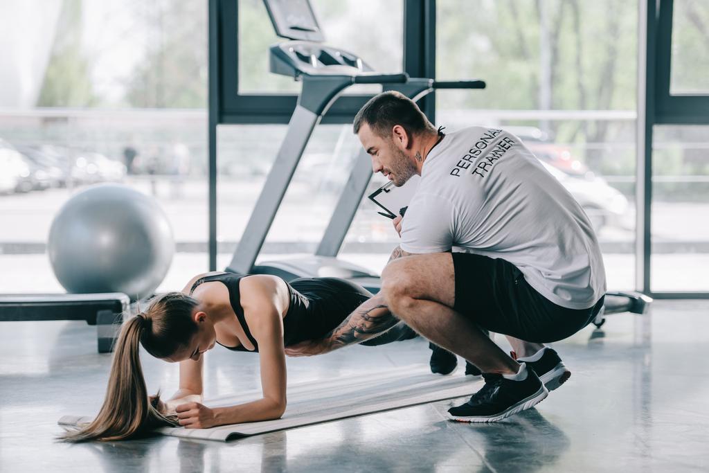 férfi személyi edző segít fiatal sportos nő deszka, fitness szőnyeg edzőteremben csinál - Fotó, kép