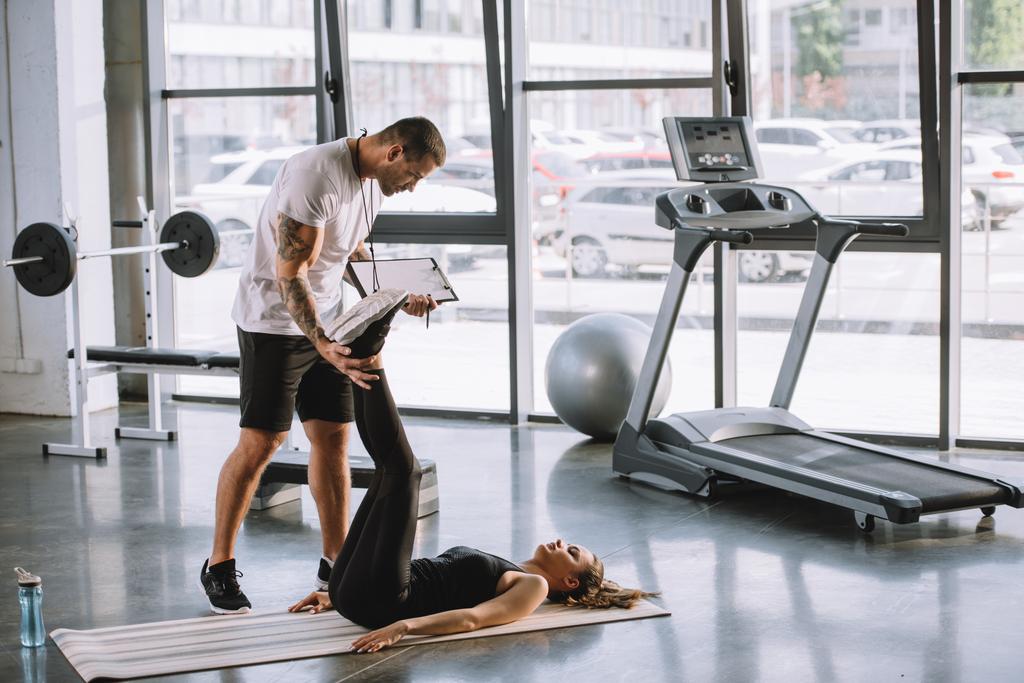mannelijke persoonlijke trainer helpen van jonge sportieve vrouw te doen abs op fitness mat op sportschool - Foto, afbeelding
