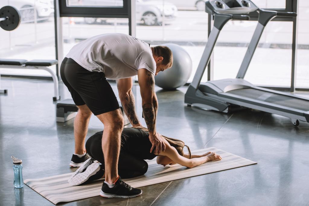 férfi személyi edző segít fiatal sportos nő stretching edzőteremben fitness szőnyeg - Fotó, kép
