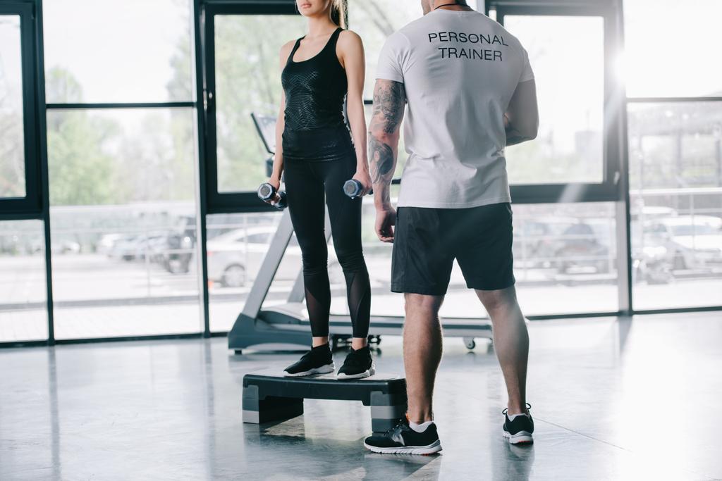 περικοπεί shot αρσενικό προσωπικό γυμναστή και νεαρό αθλήτρια που κάνει το βήμα αεροβική άσκηση με βάρη στο γυμναστήριο - Φωτογραφία, εικόνα