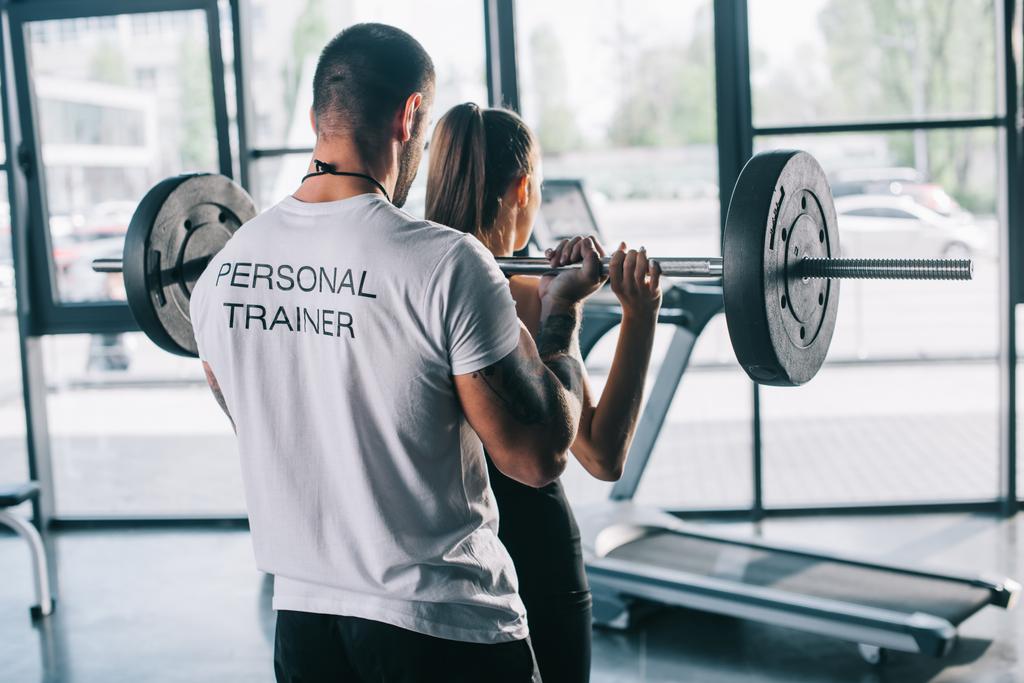 male personal trainer helping sportswoman to do exercises with barbell at gym - Photo, Image
