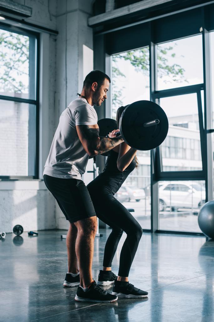 male personal trainer helping sportswoman to do exercises with barbell at gym - Photo, Image