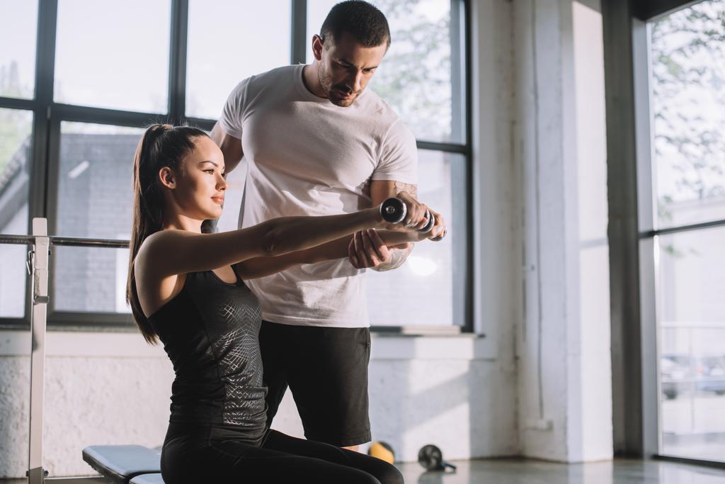 männlicher Personal Trainer hilft Sportlerin bei Übungen mit Kurzhanteln im Fitnessstudio - Foto, Bild